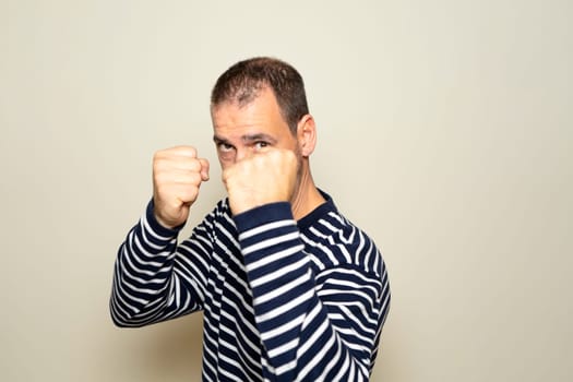 Bearded Hispanic man in his 40s wearing a striped sweater with his fists in defense position prepared to fight, isolated on beige isolated background