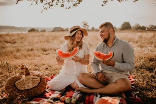 happy European caucasian family with a pregnant woman relaxing in nature picnic eating fruit red juicy watermelon laugh having fun. expectant mother in hat and dress eating watermelon in summer.