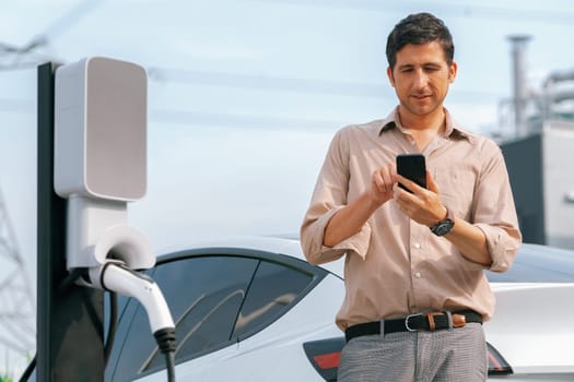 Man pay for electricity with smartphone while recharge EV car battery at charging station connected to power grid tower electrical as electrical industry for eco friendly car utilization.Expedient