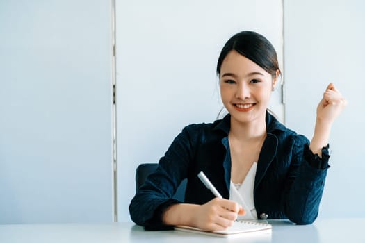 Beautiful young Asian woman writes letter on notebook while sitting at office desk. Content writer and secretary job concept. uds