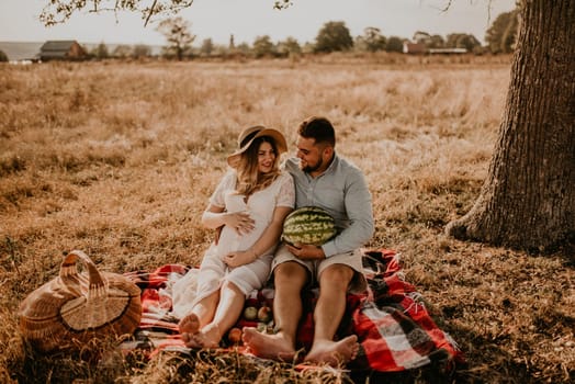 happy European caucasian family with a pregnant woman relaxing in nature picnic eating fruit red juicy watermelon laugh having fun. expectant mother in hat and dress eating watermelon in summer.