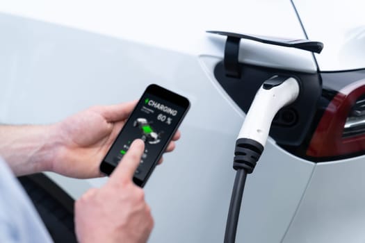 Modern eco-friendly man recharging electric vehicle from EV charging station, using Innovative EV technology utilization for tracking energy usage to optimize battery charging on smartphone.Synchronos