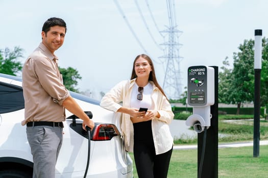 Young couple use smartphone to pay for electricity at public EV car charging station green city park. Modern environmental and sustainable urban lifestyle with EV vehicle. Expedient