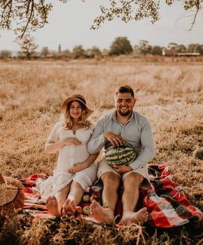 happy European caucasian family with a pregnant woman relaxing in nature picnic eating fruit red juicy watermelon laugh having fun. expectant mother in hat and dress eating watermelon in summer.