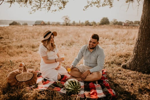 happy European caucasian family with a pregnant woman relaxing in nature picnic eating fruit red juicy watermelon laugh having fun. expectant mother in hat and dress eating watermelon in summer.