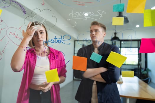 Couple of businesspeople discussing and brainstorming about solving marketing problem in front of glass board at business meeting by using mind map and sticky notes. Working together. Immaculate.