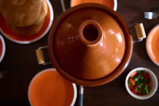 Top view clay pot tagine, freshly baked break and empty clean orange plates on wooden table. Moroccan traditional food for family dinner