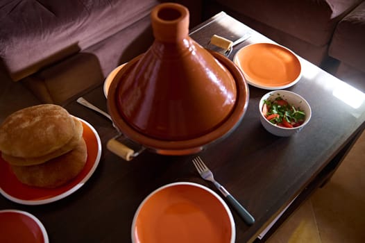 Traditional Moroccan family dinner - a delicious meal cooked in tagine clay pot with freshly baked bread on the table at home interior