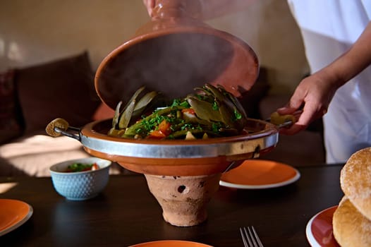 Close-up of the hands of a housewife opening the clay lid of Moroccan tagine while serving table for dinner at home