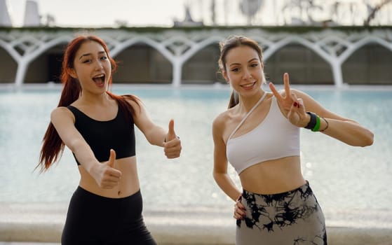 Two young female sportswomen have a rest after morning jogging outdoors and looks camera with smile