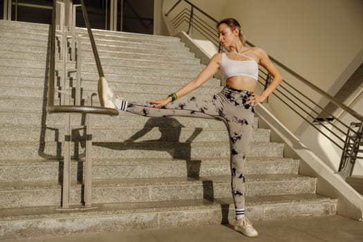 Ahletic woman in sportswear raising leg on railing and stretching during workout outside in morning