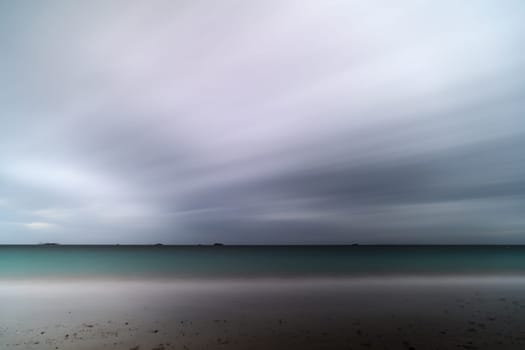 Long exposure shot of a stormy beach with clear islets on the horizon and blurred details.