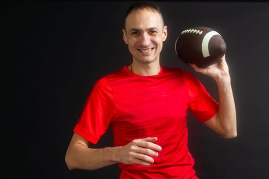 Young man dressed in casual street clothes holds a football, ready to throw a pass, isolated on black background. High quality photo