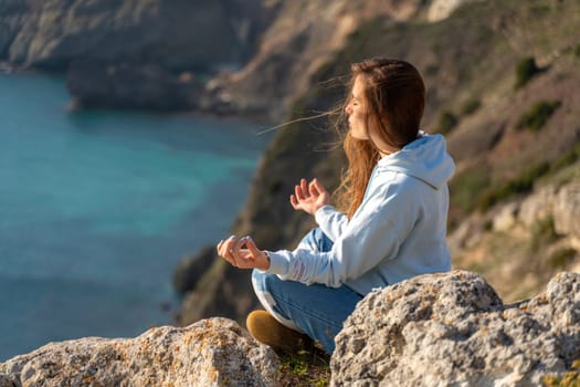 Woman tourist enjoying the sunset over the sea mountain landscape. Sits outdoors on a rock above the sea. She is wearing jeans and a blue hoodie. Healthy lifestyle, harmony and meditation.