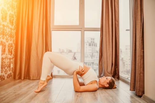 Young athletic attractive woman practicing yoga. Works out at home or in a yoga studio, sportswear, white pants and a full-length top indoors. Healthy lifestyle concept.