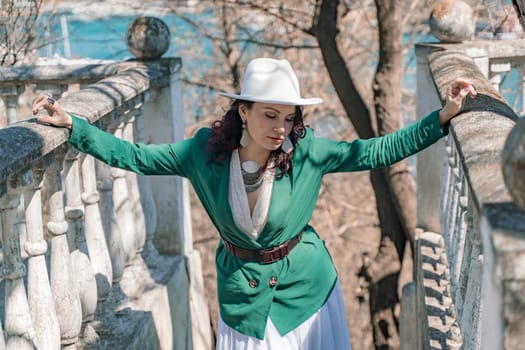 Woman walks around the city, lifestyle. Happy woman in a green jacket, white skirt and hat is sitting on a white fence with balusters overlooking the sea bay and the city