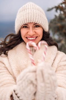 Woman candy sea. Smiling woman in knitted hat, mittens and beige coat holding lollipops candy canes in her hands in shape of heart against the backdrop of the sea