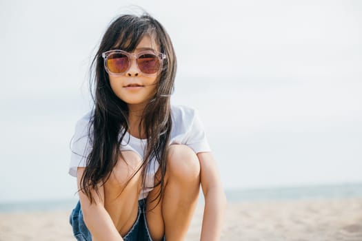 Fashionable child girl in trendy sunglasses sitting on a deck chair at the beach. Smiling portrait exuding joy and happiness. Windy hair stylish beachwear and carefree relaxation