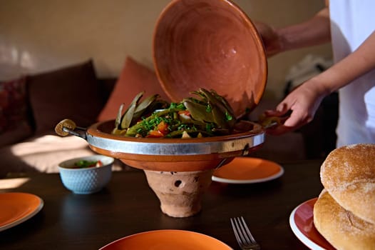 Still life. Moroccan tagine. Housewife serving delicious vegetarian meal with steamed vegetables, cooked in clay pot according to traditional Moroccan recipe. Cooked artichoke with greens and veggies