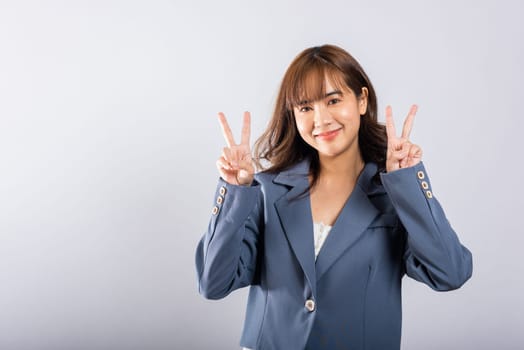 Captured in a studio with a white backdrop, a happy Asian woman marks victory and peace with her fingers near her eye. Her radiant smile and positive energy are truly contagious.