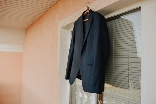 A black jacket hangs against a white window in the room. Men's dark blue brown black formal classic blazer. fashion clothing for business men