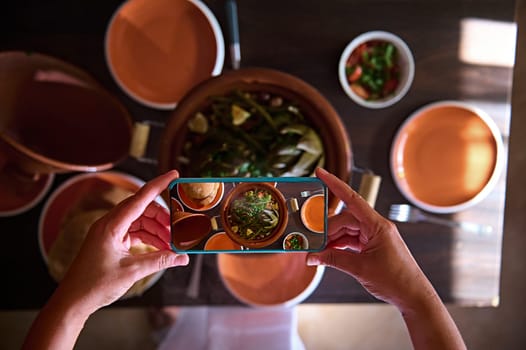 Directly above food blogger hands using smart mobile phone, taking photo of steamed veggies in clay dish tagine. Close-up of a woman photographing or recording video of Moroccan tagine for video blog