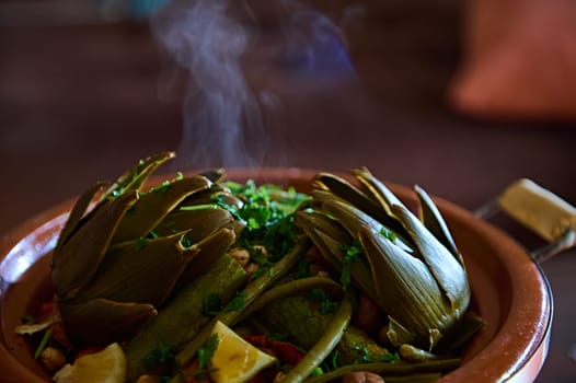 Close-up view of a hot vegetarian meal with organic veggies steamed in clay pot tagine with steam. Moroccan cuisine, culture, traditions and crockery. Extreme close-up for food blogs or advertisement