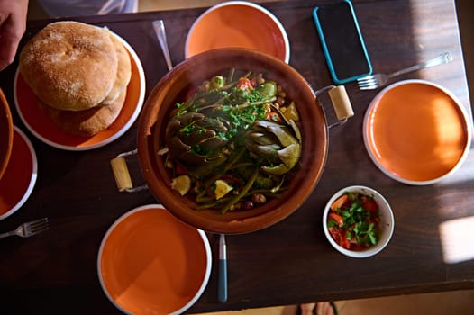 Top view wooden table served with freshly backed traditional Moroccan wholegrain bread, a tagine with delicious organic veggies with steam. Background food and culinary blogs. Copy advertising space