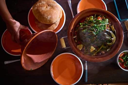 View from above of a housewife opening lid of clay crockery, showing a vegetarian meal with organic veggies steamed in clay tagine with steam. Moroccan cuisine, culture, traditions and crockery.