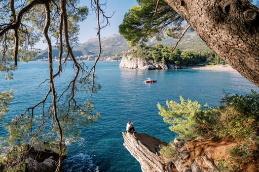 View through the green branches on a man sitting on a stone ledge and looking at a boat passing by the shore. Back view. High quality photo