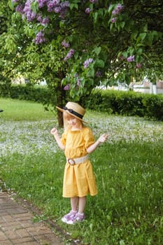 A little girl in a yellow dress and straw hat wearing a bouquet of lilacs. A walk in a spring park, blossoming lilacs