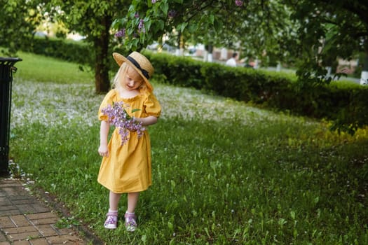 A little girl in a yellow dress and straw hat wearing a bouquet of lilacs. A walk in a spring park, blossoming lilacs