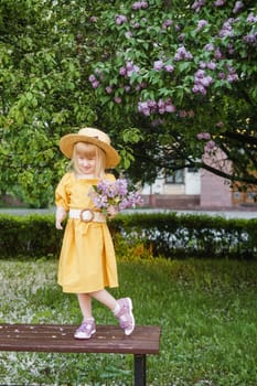 A little girl in a yellow dress and straw hat wearing a bouquet of lilacs. A walk in a spring park, blossoming lilacs