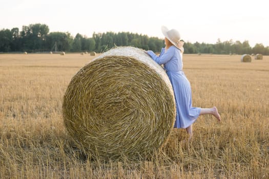 A red-haired woman in a hat and a blue dress walks in a field with haystacks