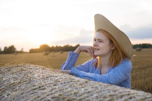 A red-haired woman in a hat and a blue dress walks in a field with haystacks