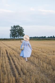 A red-haired woman in a hat and a blue dress walks in a field with haystacks