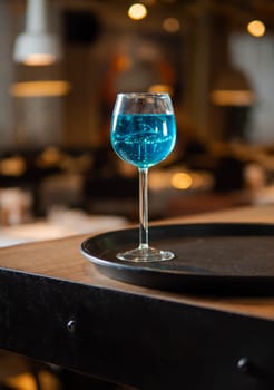 Glass of blue lagoon cocktail in shark high stem cocktail glass on beautiful bar counter background with bokeh. Aesthetic photo of blue lagoon cocktail in evening light, restaurant or bar interior
