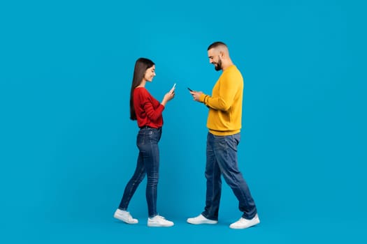 Communication, Technology And Lifestyle Concept. Smiling young couple using smartphones while walking past each other, man and woman standing isolated over blue studio background, full length