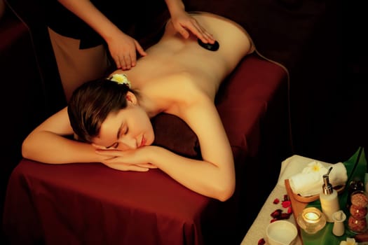 Hot stone massage at spa salon in luxury resort with warm candle light, blissful woman customer enjoying spa basalt stone massage glide over body with soothing warmth. Quiescent