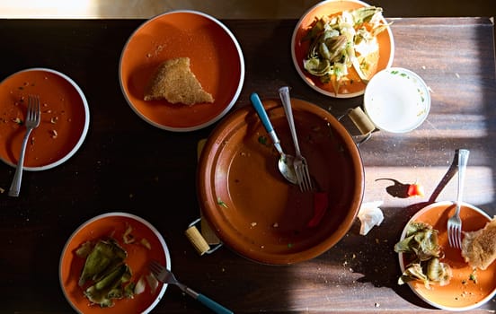 Top view of empty clay dish tagine and dirty plates with the rest of food after dinner on a wooden table. Directly above Leftover of lunch