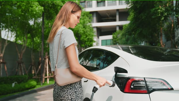 Young woman recharge EV electric car battery at residential area EV charging station in urban city condo with sustainable green and renewable clean energy lifestyle for electric vehicle innards