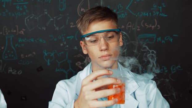 Closeup of boy inspect chemical solution while holding beaker at blackboard with chemical theory. Smart scientist doing experiment while analysis colored solution while wearing lab coat. Edification.