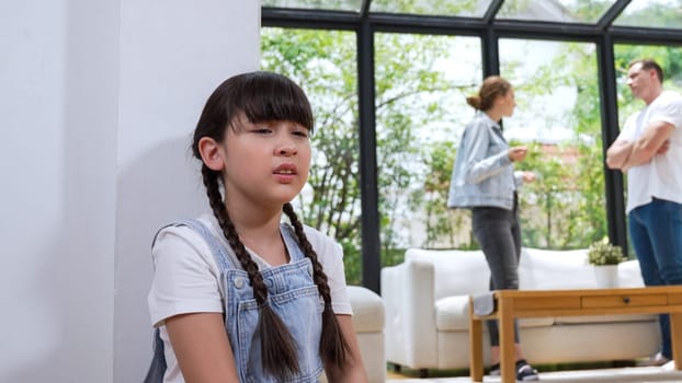 Stressed and unhappy young girl huddle in corner, cover her ears blocking sound of her parent arguing in background. Domestic violence at home and traumatic childhood develop to depression. Synchronos