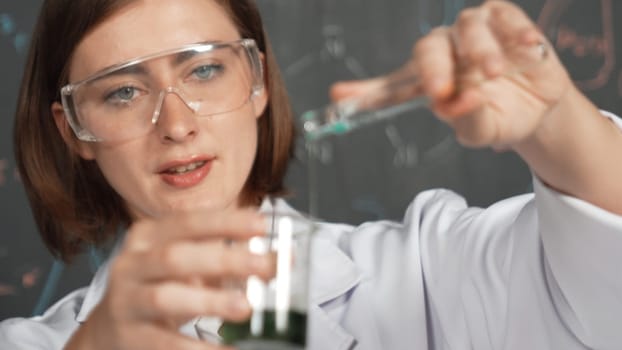 Closeup of young beautiful teacher focus on mixing chemical liquid at laboratory. Skilled scientist doing an experiment by inspect colored solution while standing in front of blackboard. Erudition.