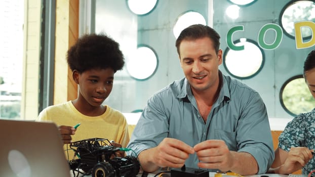 Skilled teacher teaching diverse students fixing car model by using wire and electronic equipment. Girl studying about using electrical tool to check mother board while looking at laptop. Edification