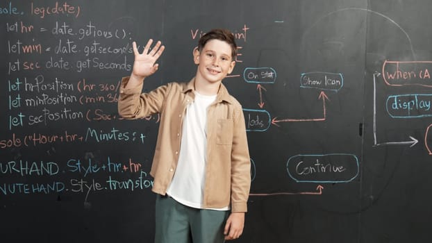 Panorama shot of happy caucasian boy smiling and waving hand at blackboard with engineering code or prompt written. Smart child looking at camera and greeting while study in STEM classroom. Erudition.