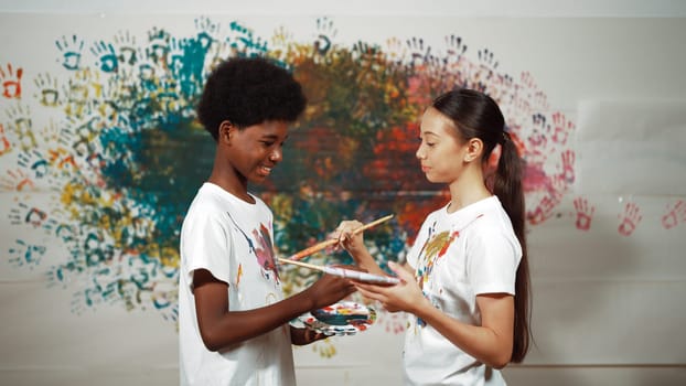 Smiling diverse children using paint brush painted color on each other white shirt shirt at colorful stained wall in art lesson. Represent exchanging experience, learning each other. Edification.