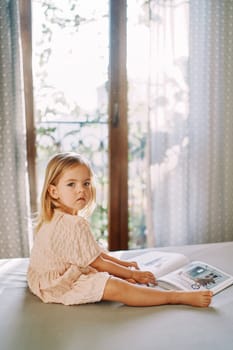 Little girl sits on a bed with a picture book and looks away. Side view. High quality photo
