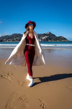 Stylish woman with maroon dress and white coat on Playa de la Concha, San Sebastian