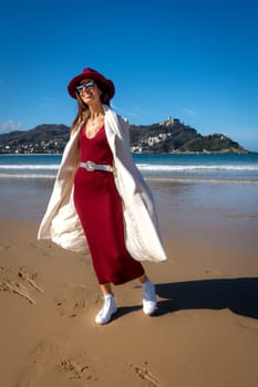 Stylish woman with maroon dress and white coat on Playa de la Concha, San Sebastian